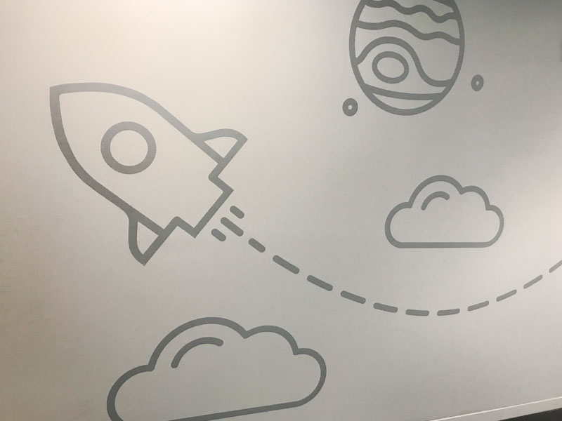 Folienbeschriftung einer weißen Wand mit hellgrauer Folie, es sind Wolken, Planeten und eine Rakete zu sehen
